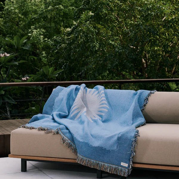 Lichtblauwe deken met franjerand en wit zonnesymbool, op outdoorbank.