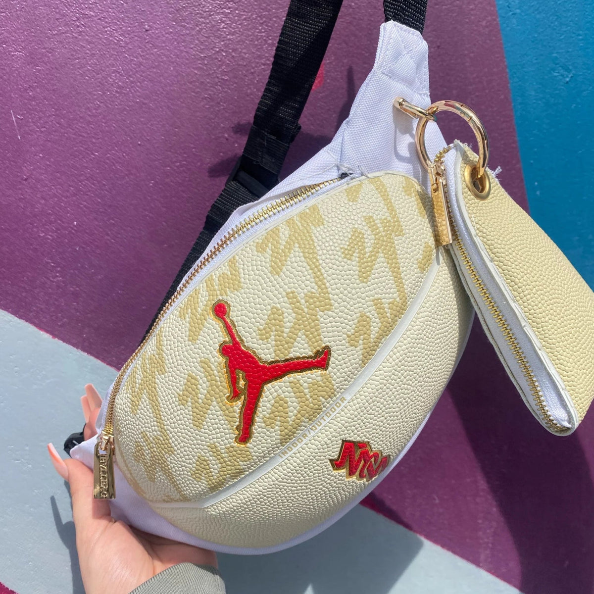 Crèmekleurige heuptas met zwarte riem en rood Jordan-logo, goudkleurige sluiting, hand vasthoudt